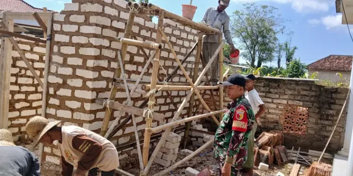 Babinsa Ganding Serma Misriadi berpakaian dinas loreng bersama warga sedang memasang batu bata dinding rumah program RTLH di Sumenep.