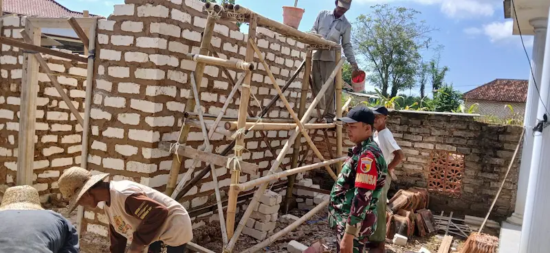Babinsa Ganding Serma Misriadi berpakaian dinas loreng bersama warga sedang memasang batu bata dinding rumah program RTLH di Sumenep.